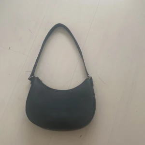 Svart axelväska i skinnlook - En stilren svart axelväska med minimalistisk design och rundad form. Väskan har ett smidigt axelband och stängs med en silvrig dragkedja.