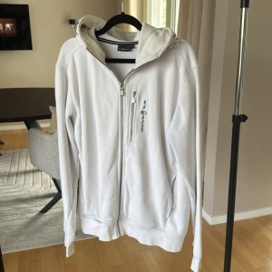 Vit hoodie från Sail Racing med huva - Vit hoodie från Sail Racing med dragkedja framtill, bröstficka med logga och justerbar huva. Tröjan har långa ärmar och två sidofickor. Perfekt för dig som gillar stilren och sportig look.