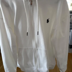  Polo Ralph Lauren - Snygg vit hoodie från Polo Ralph Lauren med dragkedja framtill, huva med snörning och klassisk broderad logga på bröstet. Tröjan har känguruficka och är tillverkad i mjuk bomull. Perfekt för en clean och stilren look.