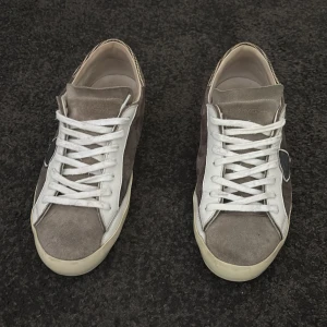 Grå mockasneakers från Philippe Model - Snygga sneakers från Philippe Model i grå mocka med vita skinnpaneler och vita snören. Skorna har en beige sula och rutigt band bak på hälen. Klassisk låg modell med rund tå och diskret logga på sidan.