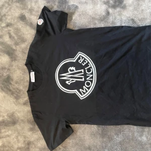 Svart Moncler t-shirt med stor logga - Svart t-shirt från Moncler med stor vit logga framtill och klassisk rund hals. På ärmen sitter en broderad Moncler-patch. T-shirten är i mjuk bomull och har en clean, stilren vibe som passar perfekt till jeans eller shorts. Storlek L passar M