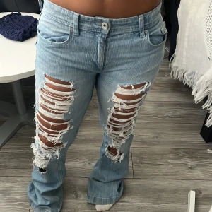 Ljusblå slitna bootcut jeans H&M - Säljer ett par ljusblå bootcut jeans från H&M med kraftiga slitningar och hål framtill på benen. Jeansen har flared ben, justerbar midja och klassiska bakfickor med knappar och slitna detaljer. Materialet är denim i bomull.