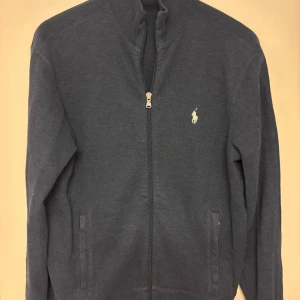 Blå kofta från Ralph Lauren - Mörkblå kofta från Ralph Lauren med dragkedja framtill och klassisk broderad logga på bröstet. Tröjan har hög krage, två fickor och är långärmad. Perfekt för lager-på-lager och chill stil.