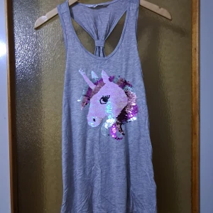 Grå linnetopp med unicorn - Grå ärmlös linnetopp med snygg knut i ryggen och stort paljettmotiv av en enhörning på framsidan. Paljetterna skiftar färg när du drar med handen, vilket ger en cool effekt. Jätte fin- använd endast en gång :) passar för 14+ år. Passar upp till storlek M/L 