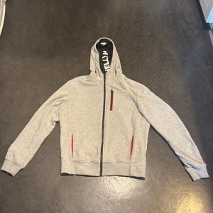 Grå Helly Hansen hoodie med dragkedja - Snygg ljusgrå hoodie från Helly Hansen med hel dragkedja och huva. Tröjan har röda detaljer vid fickorna och bröstficka med dragkedja. Mjukt material, perfekt för chill eller utomhusaktiviteter. Långärmad och med logga på axeln.