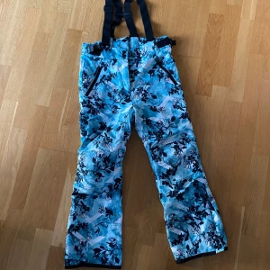 Blommiga täckbyxor från Everest 140 - Säljer ett par täckbyxor från Everest i storlek 140. De är blå och vita med blommigt mönster, har svarta hängslen och dragkedjor vid fickorna. Perfekta för vintern och håller dig varm och torr i backen.