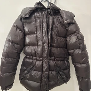 Svart Moncler dunjacka med huva - Snygg svart dunjacka från Moncler med glansig finish, avtagbar huva och flera fickor. Jackan har dragkedja framtill, logga på ärmen och quiltad design som ger en riktigt fet vinterlook. Perfekt för dig som vill hålla stilen när det är kallt ute.