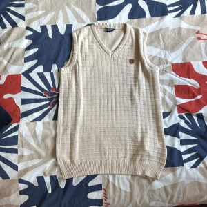 Beige stickad väst från LC Waikiki - Beige stickad väst med v-ringning och diskret rutmönster. Västen har ribbade kanter vid hals, ärmhål och nederkant samt en liten brun logodetalj på bröstet. Perfekt för lager-på-lager och en clean look.