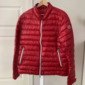 Röd dunjacka från Moncler - Säljer en röd dunjacka från Moncler med klassisk quiltad design och två dragkedjefickor framtill. Jackan har hög krage, Moncler-logga på ärmen och stängs med dragkedja. Perfekt för kyliga dagar när du vill ha både stil och värme. Pris kan diskuteras
