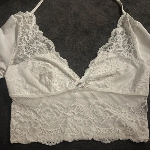 Vit spetsig croppad långärmad topp - Superfin vit croppad topp med långa ärmar och spetsdetaljer framtill och baktill. Djup v-ringning i både baksida och fram och knytband i nacken ger en snygg och feminin look. Perfekt för dig som gillar romantiska vibes och vill sticka ut lite extra.