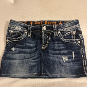 Rock Revival jeanskjol med broderi - Snygg kort jeanskjol från Rock Revival med slitna detaljer, kontrastsömmar och broderade bakfickor med nitar. Mörkblå tvätt och coola knappar med fleur-de-lis. Perfekt för dig som gillar streetstyle, Stockholmsstil och statement-plagg.