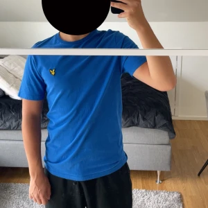 Blå t-shirt från Lyle & Scott - Snygg blå t-shirt från Lyle & Scott med klassisk rund hals och korta ärmar. På bröstet sitter den ikoniska gula örnloggan som ger en clean och sportig vibe. Tillverkad i mjuk bomull som känns skön mot huden. Perfekt till jeans eller shorts.