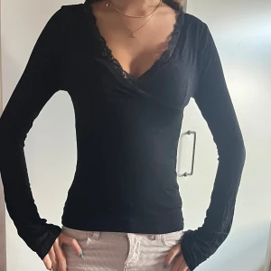 Svart omlottopp med spetsdetalj från Nelly - Snygg svart omlottopp med v-ringning och spetskant vid halsen. Långärmad och figurnära passform, perfekt att styla med jeans eller kjol. Tunn och mjuk i materialet, ger en elegant och trendig look. Jätte bra skick från Nelly