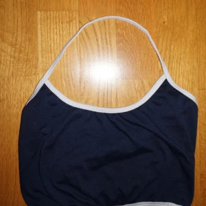 Mörkblå bandeau tubtopp med vita kanter - En stilren mörkblå bandeau tubtopp med tunna vita kantband. Toppen är ärmlös och har en enkel, sportig look som passar perfekt till sommaren. Mjuk och stretchig, troligtvis i bomullsmix. Snygg att matcha med jeans eller kjol.