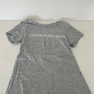 Grå t-shirt från Calvin Klein Jeans - Snygg grå t-shirt från Calvin Klein Jeans med vit logga tryckt över bröstet. Klassisk passform med rund hals och korta ärmar. Perfekt basic-plagg som funkar till allt och är superbekväm tack vare mjukt bomullsmaterial.
