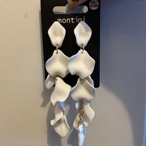 Vita örhängen från Montini - Unika vita örhängen från Montini med vågiga, blomliknande former och guldfärgade kedjedetaljer. Långa och iögonfallande, perfekta för att lyfta vilken outfit som helst. Helt oanvända!