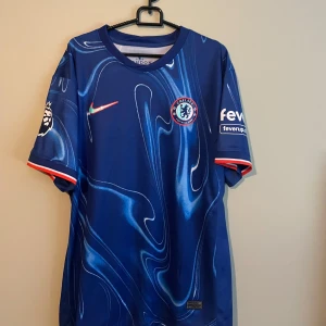 Chelsea Madueke 11 matchtröja Nike - Chelsea FC matchtröja med Madueke 11-tryck, tillverkad av lätt och ventilerande polyester. Djupblå bas med unikt swirlmönster, vita detaljer och röda inslag vid ärmslut. Premier League-logga på ärmen och klubbmärke på bröstet. Perfekt för dig som älskar fotboll och stil.