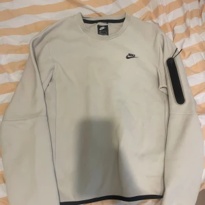Beige Nike sweatshirt med svart detalj - Säljer en stilren beige sweatshirt från Nike med svart logga på bröstet och en svart dragkedjeficka på ena ärmen. Tröjan har rund hals, långa ärmar och är tillverkad i mjuk bomullsblandning. Perfekt för en clean och sportig look.