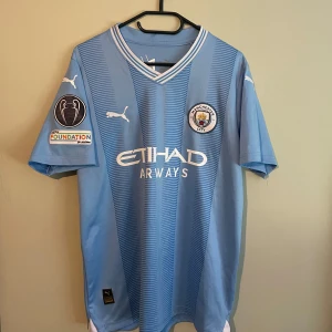 Manchester City Doku #11 matchtröja Puma - Manchester Citys officiella ljusblå matchtröja från Puma med Doku och nummer 11 på ryggen. Tröjan har korta ärmar, V-ringning och klubbmärke på bröstet. Tillverkad i lätt och ventilerande polyester, med Champions League-patch på ärmen och Etihad Airways som sponsor.