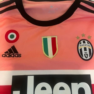 Juventus Dybala 21 rosa matchtröja - Juventus bortatröja från Adidas i rosa med svarta detaljer, Dybala 21 på ryggen och officiella Serie A och klubbmärken. Tillverkad i lätt och ventilerande climacool-material, klassisk rund hals och korta ärmar. Perfekt för dig som älskar fotboll och vill sticka ut.