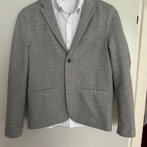 Grå Kavaj från Zara - Grå kavaj-aktig kofta i nyskick. Den är slim fit och har en enkel knapp framtill samt två knappar vid ärmsluten. Perfekt för både bal, fest och vardag. Kavajen är tillverkad i ett mjukt och bekvämt material, troligtvis bomull. Passar bra till många tillfällen! 