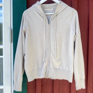 Beige zipup hoodie från 157 - Beige hoodie från 157 med dragkedja framtill och huva med snörning. Tröjan är i mjuk bomullsmix, har långa ärmar och ribbade muddar vid ärmslut och nederkant. 