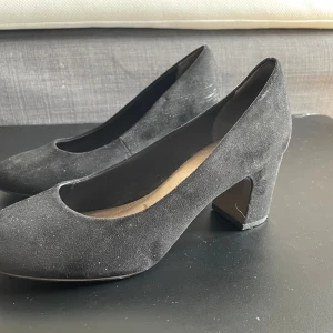 Svarta mockapumps från Tamaris - Klassiska svarta pumps från Tamaris i mjuk mocka med rund tå och stabil klack. Snygg och tidlös modell som passar till många olika outfits. Skorna har en låg klack och är bekväma att gå i tack vare den breda klacken.