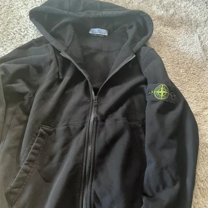 Svart Zip hoodie Stone Island - Svart hoodiejacka från Stone Island med dragkedja framtill och klassisk logotyp på ärmen. Jackan har huva med dragsko och två fickor framtill. Tillverkad i mjuk bomull för en skön och avslappnad stil. Skriv för Qr-kod