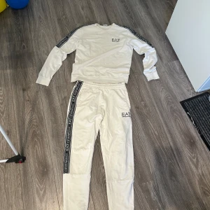 Vit EA7 Emporio Armani sweatshirt - Vit långärmad sweatshirt från EA7 Emporio Armani med svarta logoband längs ärmarna och diskret logga på bröstet. Tröjan har rund halsringning och är tillverkad i mjuk bomull, perfekt för en clean och sportig look.