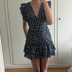 Marinblå prickig klänning - Supersöt kort mörkblå klänning med vita prickar i polka dot mönster. V-ringning och volangdetaljer vid ärmar och kjol. Öppen rygg med knytband bak ger en extra snygg touch. Endast använd en gång. Superfin och trendig nu till sensommaren/hösten!