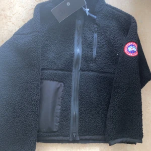 Svart teddyjacka från Canada Goose - Svart teddyjacka från Canada Goose med klassisk logga på ärmen. Jackan har hel dragkedja framtill, bröstficka med dragkedja och detaljer i nylon. Perfekt för kalla dagar och riktigt skön att bära. Snygg, enkel och stilren design.