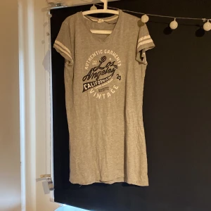 Grå t-shirtklänning Female Passion - Sportig grå t-shirtklänning från Female Passion med tryck 'Los Angeles California Vintage' framtill. Kort ärm med vita ränder, v-ringad hals och avslappnad passform. Perfekt för chill dagar eller när du vill ha en casual look.