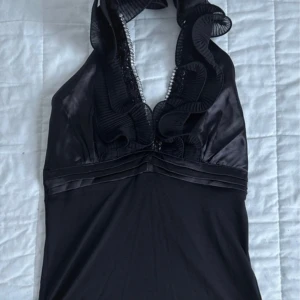 Svart halterneck topp med volang - Supersnygg svart halterneck topp med djup v-ringning, volangkant och glittriga pärldetaljer längs urringningen. Sidenliknande material som ger en lyxig känsla. Perfekt för dig som vill sticka ut med en elegant och cool look.