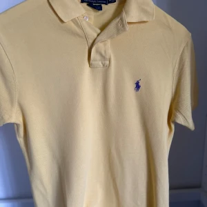 Gul pikétröja från Ralph Lauren - Snygg gul pikétröja från Ralph Lauren med klassisk krage och två knappar. Tröjan har korta ärmar och ikonisk lila broderad logga på bröstet. Perfekt för dig som gillar stilren och tidlös design. Tillverkad i mjuk bomull som andas. Perfekt till dig som vill börja resella eller ha märkeskläder till feta priser!
