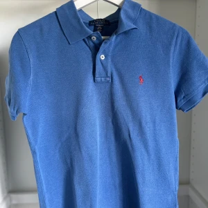 Blå pikétröja från Ralph Lauren - Klassisk blå pikétröja från Ralph Lauren med röd broderad logga på bröstet. Tröjan har korta ärmar, krage och två knappar framtill. Tillverkad i bomull och har en snygg, tidlös look som passar perfekt till jeans eller shorts. Perfekt till dig som vill börja resella eller ha märkeskläder till feta priser!
