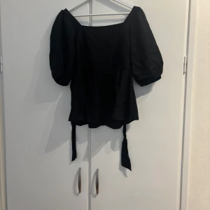 Svart blus med puffärm från AWARE - Supersnygg svart blus från AWARE med puffiga korta ärmar och knytband i ryggen. Använd vid ett tillfälle i storlek S