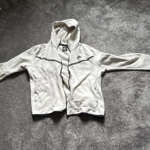 Vit Nike tech med dragkedja L - Snygg vit Nike tech från Nike i storlek L. Jackan har hel dragkedja, huva och svarta detaljer längs bröstet och dragkedjan. Tillverkad i mjukt bomullsmaterial, perfekt för chill eller träning. Klassisk Nike-logga på bröstet och två fickor framtill. Skön och varm perfekt till nu hösten och vintern, säljer för jag ej använder den mer