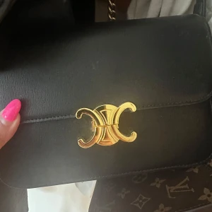 Svart axelväska i skinn - Snygg svart axelväska  i slätt skinn med ikoniskt guldspänne framtill. Väskan har justerbar axelrem med guldfärgat spänne och flera fack inuti, inklusive dragkedjefack. 