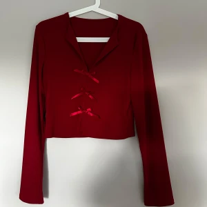 Röd croppad kofta med rosetter - Superfin röd croppad kofta med långa ärmar och snygg krage. Framsidan har tre dekorativa rosetter som knytning, vilket ger en unik och trendig look. Perfekt att styla med jeans eller kjol för en cool vibe.