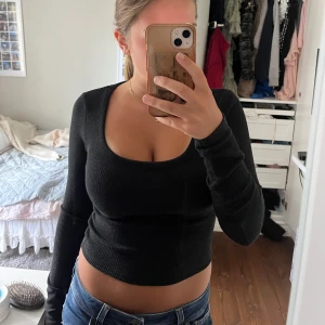 Svart ribbad långärmad topp - Säljer en svart, ribbad långärmad topp med djup rundad urringning. Modellen är croppad och sitter tight mot kroppen, perfekt till jeans eller kjol. Tidlös och enkel att styla för olika looks.