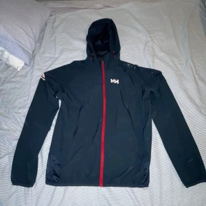 Mörkblå Windbreaker från Helly Hansen  - Svart windbreaker från Helly Hansen med huva och röd dragkedja framtill. Perfekt allvädersjacka i storlek S. Är som i nyskick och passar runt 170-180cm.