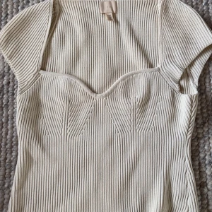 Beige ribbad topp från H&M - Säljer en beige ribbad topp från H&M med korta ärmar och markerad sweetheart-urringning. Toppen har en figurnära passform och är stickad i ett mjukt material som känns både trendigt och bekvämt. Perfekt att styla med jeans eller kjol för en snygg look.