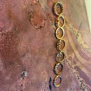 Set med sex guldiga ringar med stenar - Säljer ett set med sex snygga guldiga ringar i olika designer. Några av ringarna har glittrande stenar och andra är släta eller har mönster. Perfekt för att mixa och matcha på fingrarna för en trendig look.