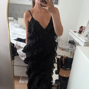 Svart långklänning med volanger - Supersnygg svart långklänning med tunna axelband och djup v-ringning. Köpt för ca 1000kr och endast använd en gång. 🖤