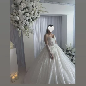 Bröllopsklänning  - Magisk vit balklänning med glittrande tyg, pärlbroderad överdel och hjärtformad urringning. Klänningen har en stor, svepande kjol och är axelbandslös. Matchande långa handskar med pärlor ingår. Perfekt för dig som vill känna dig som en prinsessa.