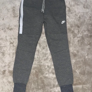 Grå mjukisbyxor från Nike - Mörkgrå mjukisbyxor från Nike med vit logga på låret, dragsko i midjan och ribbade muddar vid bensluten. Byxorna har en vit rand längs ena benet och är tillverkade i mjukt material som passar perfekt för chill eller träning.