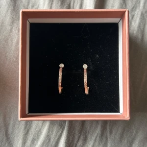 Glittriga roséguldiga örhängen - Snygga örhängen i roséguld med glittrig yta och en liten klar sten upptill. De är halvcirkelformade.