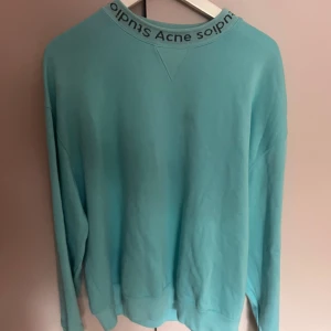 Turkos sweatshirt från Acne Studios - Säljer en turkos sweatshirt från Acne Studios med logga tryckt runt halsringningen. Tröjan har en relaxed passform, långa ärmar och är tillverkad i mjuk bomull. Perfekt för dig som gillar stilrena och cleana plagg med en twist. Tröjan har däremot ett litet hål på höger armen.