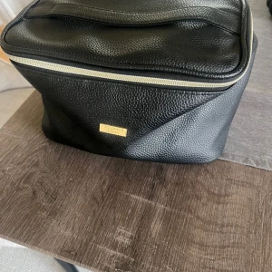 Svart necessär från Voyage - Snygg svart necessär från Voyage i skinnimitation med guldfärgad logga framtill. Väskan har en praktisk dragkedja upptill och en stilren, enkel design som passar perfekt för att organisera dina grejer när du är på språng.