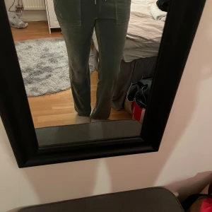 Gröna velourbyxor från Juicy Couture - Mysiga gröna mjukisbyxor från Juicy Couture i velour med broderad JC-logga framtill. Byxorna har dragsko i midjan, raka ben och fickor både fram och bak med snygga detaljer. Perfekta för chill eller streetstyle.